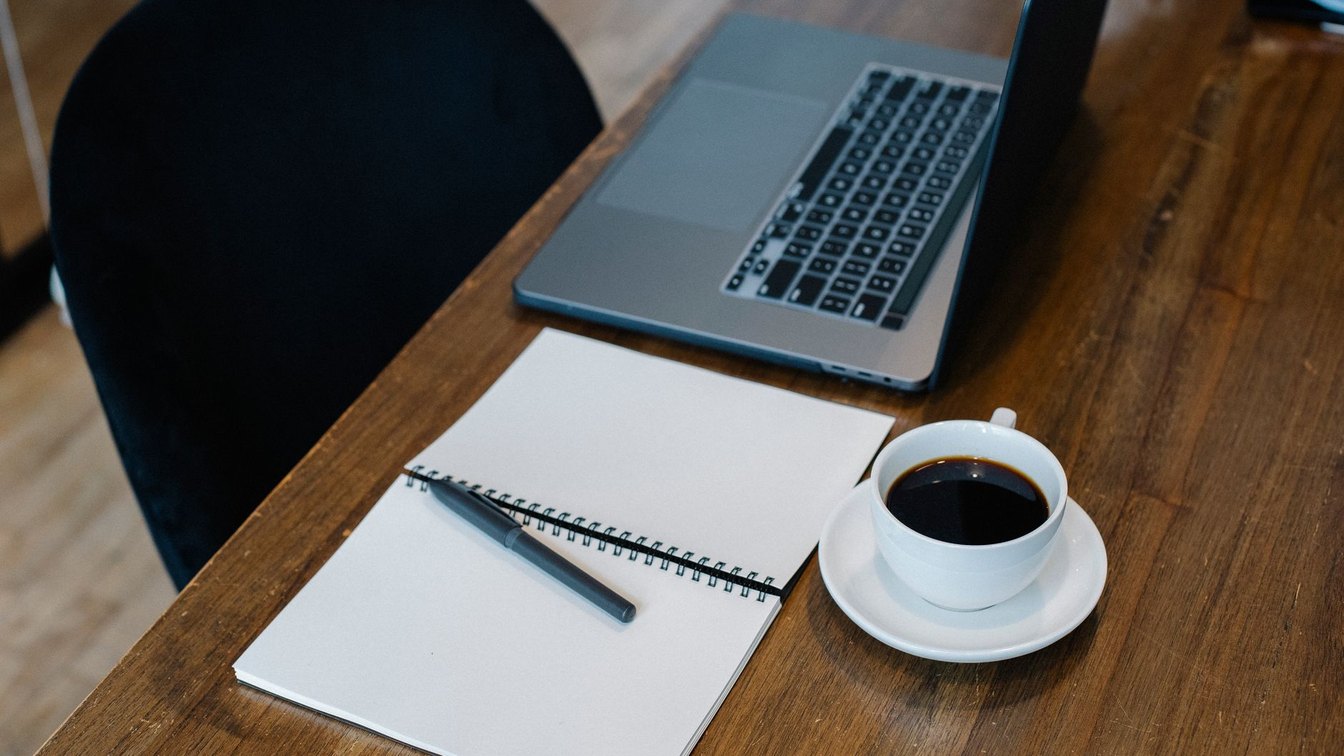 An open laptop, a spiral notebook, a pen, and a cup of coffee on a wooden desk