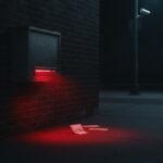 A dark night-deposit drop box on a brick wall glowing with an alarming red light from inside, envelopes scattered on the ground beneath it