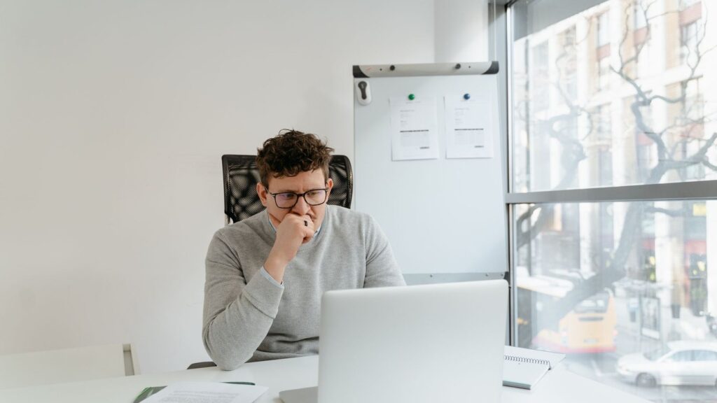 A thoughtful developer in glasses looking at a laptop screen in a bright office