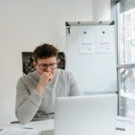A thoughtful developer in glasses looking at a laptop screen in a bright office