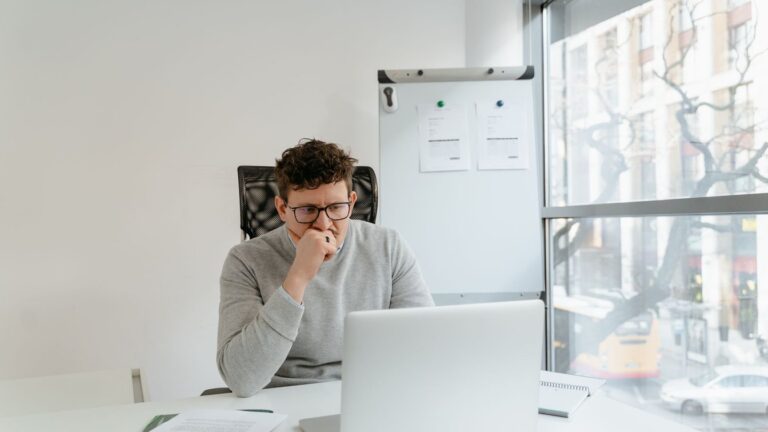 A thoughtful developer in glasses looking at a laptop screen in a bright office