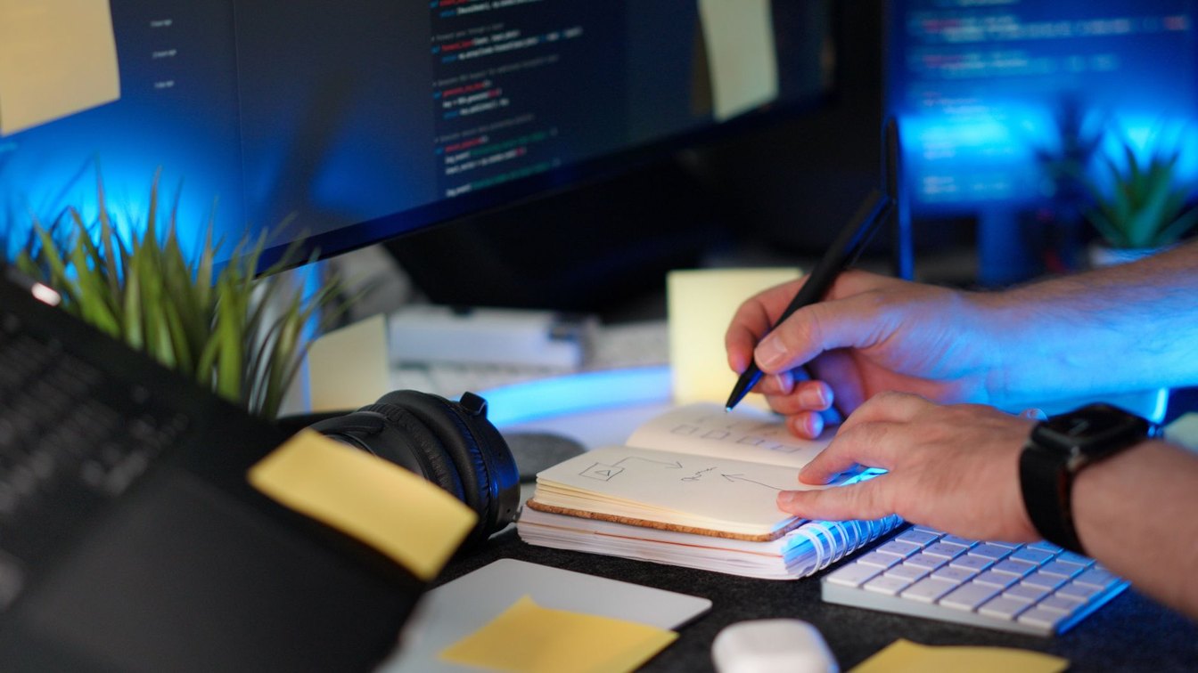 A developer sketching a checklist by hand at a desk with a code editor open on the monitor behind them