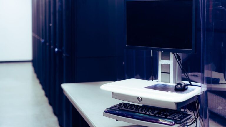 Empty terminal desk facing a row of dark server racks lit in blue, in a data center corridor