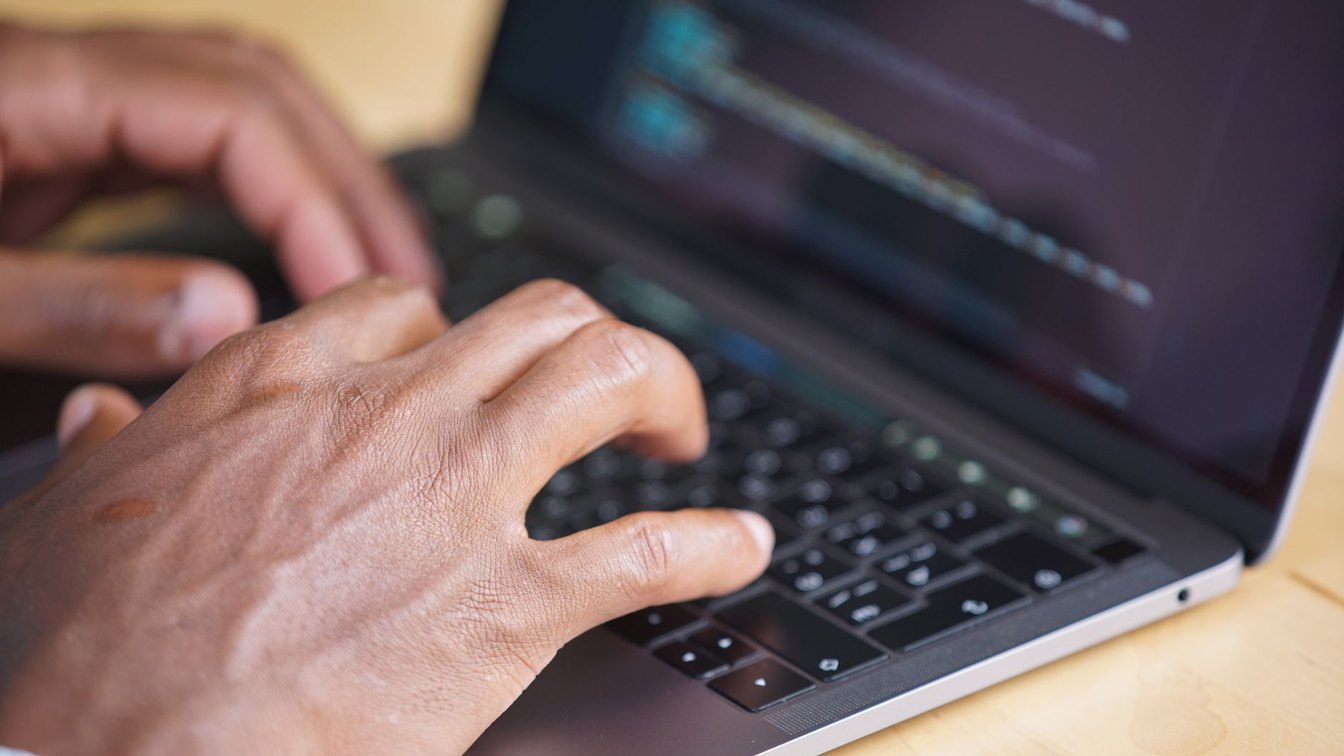Close-up of hands typing on a laptop keyboard with code visible on the blurred screen