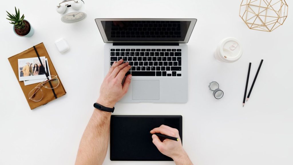 Overhead view of a designer's desk with a laptop, a drawing tablet and stylus, coffee, and notebooks