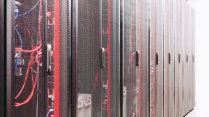 A dim data center aisle at night with rows of server racks, one rack unit glowing an alarm-red color among the others