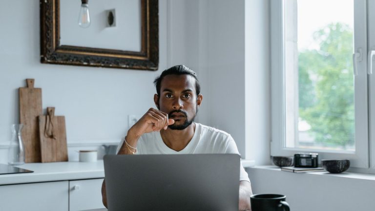 A person sitting at a laptop in a bright, minimalist home office, thinking
