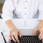 Close-up of a person's hands typing on a laptop keyboard in a bright, modern office